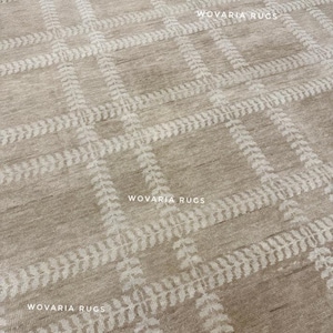 May include: Beige area rug featuring a repeating grid pattern. The grid is formed by white, leaf-like designs. The rug has a textured appearance and the text "WOVARIA RUGS" is visible in multiple places.