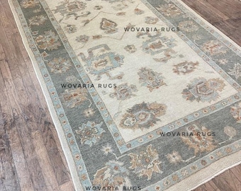 May include: Rectangular rug with a cream-colored central field and a border in shades of gray and blue. The rug features floral patterns and the text "WOVARIA RUGS" in multiple locations. The rug is placed on a wooden floor.