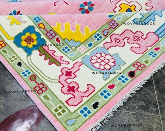 May include: A rectangular area rug featuring a pink central field with a white border. The design includes geometric floral motifs in blue, yellow, green, and red, with the text WOVARI RUGS repeated across the surface and white fringe edges.