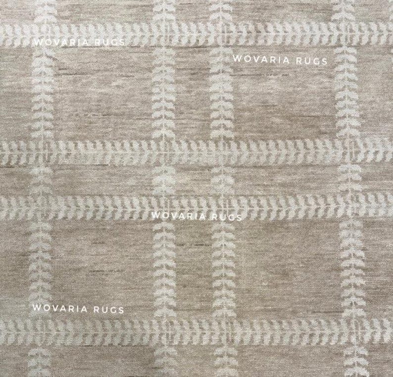 May include: A beige rug with a grid pattern. The design incorporates vertical and horizontal lines with a repeating leaf-like motif in a lighter shade. The text "WOVARIA RUGS" is printed across the image.