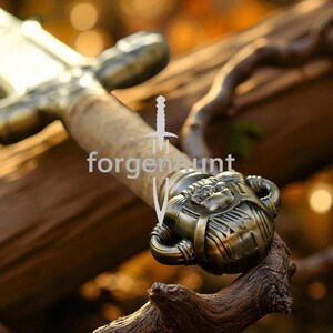 May include: Close-up of a sword with a bronze-colored hilt and a light-colored grip. The hilt features intricate detailing and a decorative design. The text "forgenhunt" is visible on the image. The sword is resting on a wooden branch.