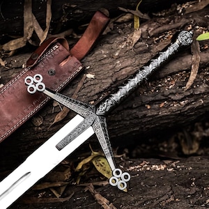 Pode incluir: Espada medieval com punho e guarda prateados e lâmina de dois gumes. A espada está sobre uma superfície de madeira, ao lado de uma bainha de couro castanho.