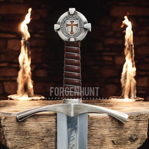 May include: A medieval-style sword with a silver blade and crossguard. The handle is wrapped in brown leather. The pommel features a shield with a cross design. The word "FORGENHUNT" is visible on the wooden surface.