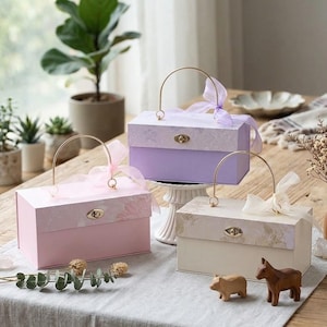 May include: Three decorative gift boxes in pink, lavender, and cream, each with a gold handle and a matching ribbon. The boxes have a floral pattern on the lid and a gold clasp. A wooden pig and horse are on the table.