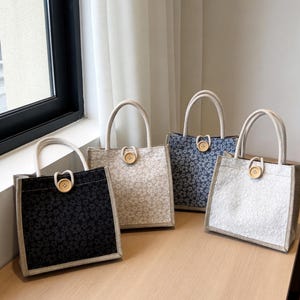 May include: Four floral tote bags with rope handles and wooden button closures. The bags feature different colors: black, beige, blue, and white, each with a floral pattern. The bags are displayed on a wooden surface.