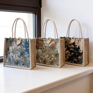 May include: Three tote bags with floral tapestry designs. Each bag has a natural jute body, rope handles, and a wooden button closure. The floral patterns feature shades of blue, green, brown, and black.