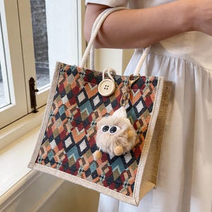 May include: A square tote bag with a geometric pattern in shades of red, orange, teal, and brown. The bag has rope handles and a small, fluffy keychain. A large button is attached to the bag's front.