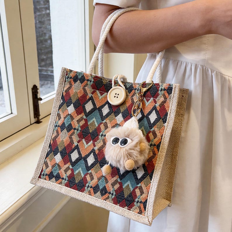 May include: A square tote bag with a geometric pattern in shades of red, orange, teal, and brown. The bag has rope handles and a small, fluffy keychain. A large button is attached to the bag's front.