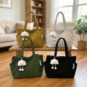Puede incluir: Cuatro bolsos tote de pana en color amarillo mostaza, gris claro, verde oliva y negro. Cada bolso cuenta con dos asas superiores, bolsillos laterales con cierre de cord&oacute;n y un dije de nube blanca con pompones colgantes en el frente.