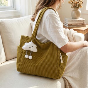 May include: Olive green corduroy tote bag with a cloud-shaped charm. The bag has two exterior pockets and long shoulder straps. The cloud charm has a smiling face and dangling pom-poms. The bag is a stylish accessory.