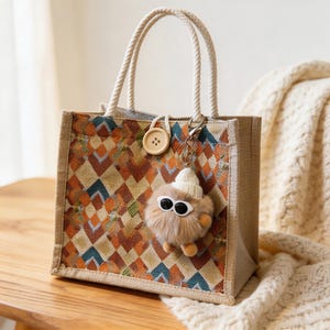 May include: A small tote bag with a geometric pattern in shades of orange, brown, and blue. The bag has rope handles and a decorative button. A fluffy keychain with sunglasses and a small hat hangs from the bag.