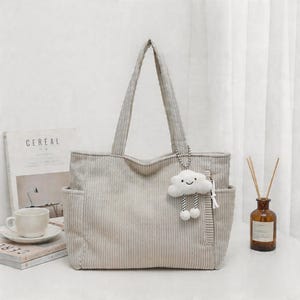 Puede incluir: Un bolso tote de pana en color beige claro con dos correas para el hombro, bolsillos laterales y un compartimento vertical con cremallera. Un dije de peluche blanco con forma de nube, cara sonriente y piernas colgantes est&aacute; sujeto al bolso.