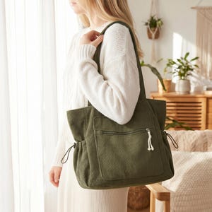 Puede incluir: Un bolso tote de pana verde oliva que presenta un gran bolsillo exterior frontal con cierre de cremallera horizontal, un tirador de cremallera de cordón blanco anudado y cordones laterales ajustables con finas cuerdas negras.
