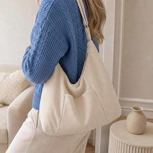 May include: A cream-colored corduroy hobo bag featuring a wide shoulder strap, a zippered top closure, and a single exterior slip pocket on the front. The bag is made of a textured, ribbed fabric and has a slouchy, unstructured shape.