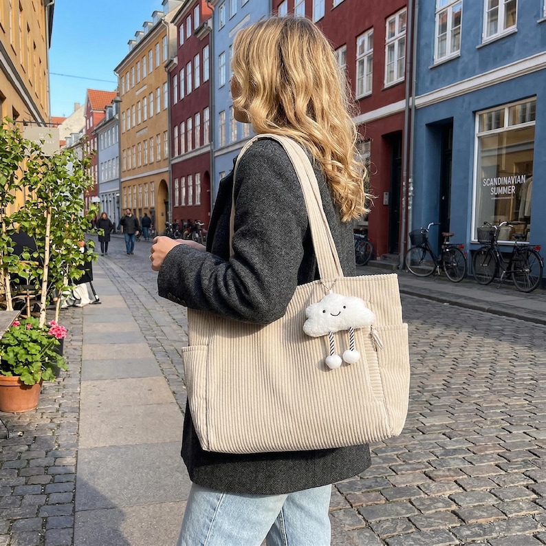Puede incluir: Un bolso tote de pana beige con dos correas para el hombro y bolsillos laterales. Un peque&ntilde;o dije de peluche blanco con forma de nube, cara sonriente y dos piernas colgantes con pies redondos est&aacute; sujeto al frente del bolso con un clip plateado.