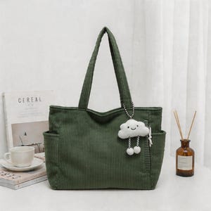 Puede incluir: Un bolso tote de pana en color verde oliva oscuro con dos correas para el hombro, bolsillos laterales y un llavero de peluche blanco con forma de nube, cara sonriente y piernas colgantes sujeto por una cadena de cuentas plateada.