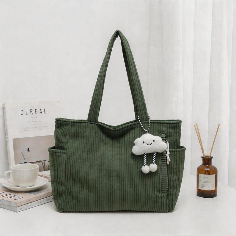 Puede incluir: Un bolso tote de pana en color verde oliva oscuro con dos correas para el hombro, bolsillos laterales y un llavero de peluche blanco con forma de nube, cara sonriente y piernas colgantes sujeto por una cadena de cuentas plateada.