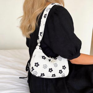 Könnte beinhalten: Weiße Umhängetasche mit schwarzem Blumenmuster. Die Tasche hat eine geschwungene Form und einen passenden Riemen mit dem gleichen Blumenmuster. Die Tasche ist aus geripptem Stoff gefertigt und hat einen silberfarbenen Reißverschluss.