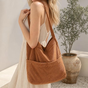 Puede incluir: Un bolso tote de pana color óxido con una textura suave y acanalada, que presenta un bolsillo exterior grande y profundo en la parte frontal. El bolso cuenta con una sola correa ancha para el hombro y un diseño espacioso y flexible.