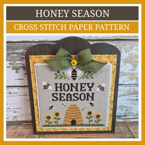 May include: A cross stitch pattern titled Honey Season featuring a central beehive, sunflowers, and bees on a light gray fabric. The design is mounted on a dark wooden board with a yellow honeycomb border and topped with a green fabric bow.