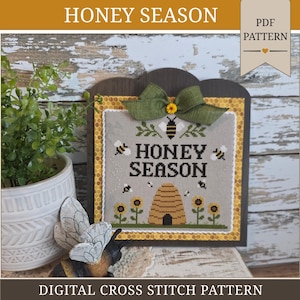 May include: A cross stitch pattern featuring the text HONEY SEASON in black, surrounded by embroidered bees, sunflowers, and a beehive on a light grey fabric. The design is framed in a dark wooden board with a yellow honeycomb border and a green bow.