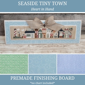 May include: A premade finishing board featuring a cross-stitch design of a seaside town with a lighthouse, houses, and the text "sea". The board is framed in light blue and adorned with a tan bow. The text "SEASIDE TINY TOWN" and "Heart in Hand" are visible.