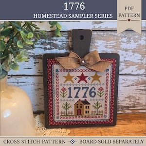May include: A framed cross-stitch sampler with the year "1776" in blue, featuring stars, a house, and floral elements. The sampler is mounted on a dark wooden board with a burlap bow. The text "HOMESTEAD SAMPLER SERIES" is visible.