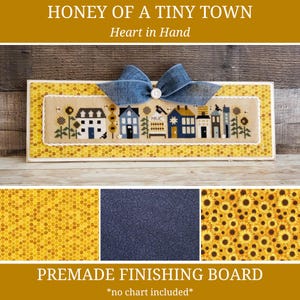 May include: A premade finishing board featuring a cross-stitch design of a tiny town with houses, sunflowers, and the text "HONEY OF A TINY TOWN". The board is decorated with a blue bow and the text "Heart in Hand".