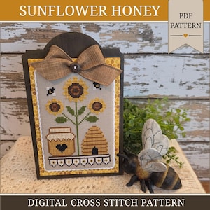 May include: A digital cross stitch pattern titled SUNFLOWER HONEY featuring a rectangular design with three sunflowers, a honey jar with a heart, and a beehive. The piece is mounted on a dark frame with a brown burlap bow and a white rope border.
