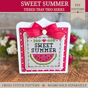 May include: A square cross-stitch piece featuring the text SWEET SUMMER in black letters above a watermelon slice graphic. The piece is mounted on a red and white patterned board with a red fabric bow and a small white flower at the top center.