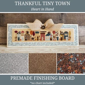 May include: A premade finishing board featuring a cross-stitch design of a tiny town with houses and trees. The board has a light blue patterned border and a burlap bow. The text "THANKFUL TINY TOWN" and "Heart in Hand" are visible.