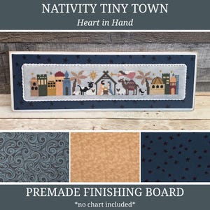 May include: A framed cross-stitch depicting a nativity scene with buildings, animals, and figures. The frame is navy blue with star accents. The text "NATIVITY TINY TOWN" and "Heart in Hand" are at the top. The bottom text reads "PREMADE FINISHING BOARD *no chart included*."