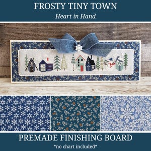 May include: A premade finishing board featuring a cross-stitch design of a winter village with houses, trees, and snowmen. The board is framed with a blue floral fabric and adorned with a blue bow and snowflake. The text "Frosty Tiny Town" and "Heart in Hand" are displayed.