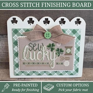 May include: A white wooden cross stitch finishing board with a scalloped top featuring shamrock cutouts. It displays a beige fabric panel with the cross-stitched text "SEW lucky," a green shamrock, a spool, scissors, and a button, topped with a tan bow.