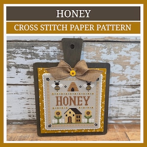 May include: A cross stitch pattern titled HONEY featuring a bee hive, two bees, a house, and sunflowers on a light gray fabric. The piece is mounted on a dark wooden board with a burlap bow and a small yellow flower accent at the top center.