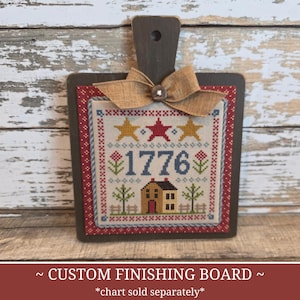 May include: A wooden custom finishing board with a stitched design featuring stars, the number 1776, and a house. The board has a dark brown handle and frame, with a burlap bow and button accent. The text "CUSTOM FINISHING BOARD" is at the bottom.