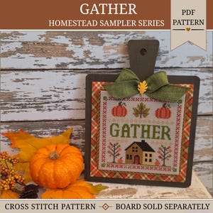 May include: A cross-stitch pattern titled GATHER HOMESTEAD SAMPLER SERIES featuring a house, pumpkins, and leaves. The square fabric is mounted on a dark wooden board with a green ribbon bow and a small yellow leaf accent on top.