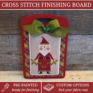 May include: A red wooden cross stitch finishing board featuring a centered cross stitch of Santa Claus holding a garland. The board is decorated with a green fabric bow and a plaid fabric border. The text CROSS STITCH FINISHING BOARD is visible.