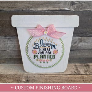 以下が含まれることがあります： 白い木製の植木鉢型のカスタム仕上げボード。ステッチデザインは、「Bloom Where You Are Planted」の文字を花柄で表現し、ピンクのドット柄のリボンでアクセントを加えています。「CUSTOM FINISHING BOARD」の文字が下部にあります。