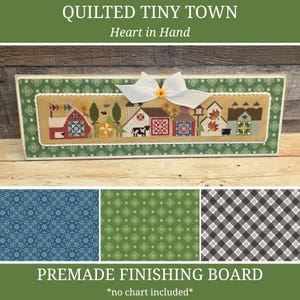 Puede incluir: Un tablero de acabado prefabricado con un diseño de punto de cruz de un pequeño pueblo con casas, un granero y una vaca. El tablero está enmarcado con un borde verde y un lazo blanco con un centro amarillo. El texto "QUILTED TINY TOWN" y "Heart in Hand" están en la parte superior.