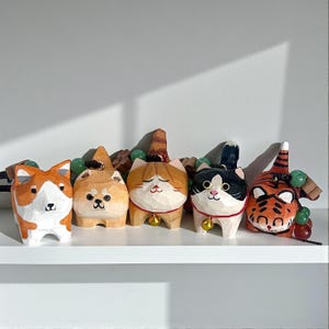 Peut inclure: Cinq figurines d'animaux en bois sculpté, dont un corgi, un Shiba Inu et des chats. Chaque figurine est ornée d'un collier à clochette et présente des détails uniques. Le tigre a des rayures orange et noires. Collection décorative sur une étagère blanche.