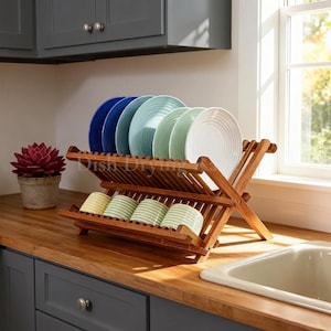 May include: A two-tier bamboo dish drying rack holding a row of colorful plates in blue, teal, and white on the top shelf and four striped mugs in yellow, green, and white on the bottom shelf, featuring a foldable X-frame construction.