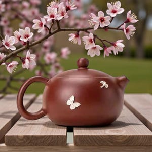 Pode incluir: Uma chaleira de cerâmica marrom com corpo arredondado, alça curva e bico pequeno. Duas decorações de borboletas prateadas adornam a chaleira. A chaleira está sobre uma superfície de madeira, com um ramo florido de flores rosas ao fundo.