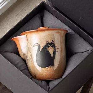May include: A ceramic teapot featuring a black cat design. The teapot has a tan and brown colour scheme, a lid, and a spout. The cat has yellow eyes and whiskers. The teapot is presented in a black gift box with grey fabric.