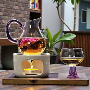 May include: A glass teapot with a purple handle and a spout, filled with tea and flowers, sits atop a ceramic warmer with a lit candle. A matching glass with a purple base is next to it on a wooden tray. The tea set is on a wooden table.