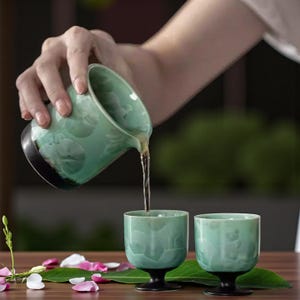 May include: A person pouring tea from a celadon-colored ceramic pitcher into two matching cups. The pitcher and cups have a textured surface and black bases. The tea is a light amber color. The scene is set on a wooden table with scattered petals and leaves.