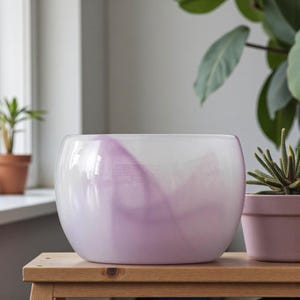 May include: A clear glass planter with a soft, rounded shape. The planter features a swirl of lavender and white, creating a marbled effect. It sits on a wooden surface, with a small potted plant visible in the background.
