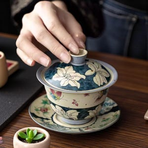 May include: A decorative tea set with a floral design. The set includes a teacup, saucer, and lid. The lid is dark blue with white floral patterns. The teacup and saucer have a cream-colored base with floral and butterfly accents.