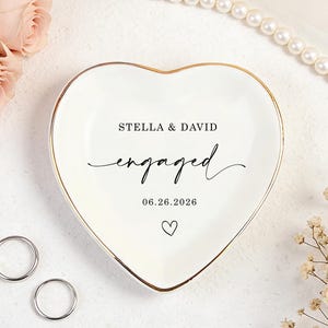 Puede incluir: Un plato de cerámica blanco en forma de corazón con borde dorado. La inscripción dice "STELLA & DAVID engaged 06.26.2026" y un símbolo de corazón. Dos anillos de plata junto al plato, con rosas rosas y perlas en el fondo.
