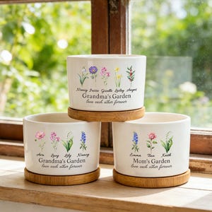 Könnte beinhalten: Drei weiße Keramik-Blumentöpfe mit floralen Designs und Bambus-Untersetzern. Jeder Topf hat eine andere Aufschrift: "Grandma's Garden" und "Mom's Garden", mit Namen und dem Satz "love each other forever".